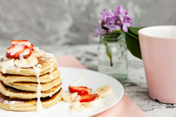 A stack of pancakes with a pink tea mug and  fresh strawberries on a white plate on a light background. A small bouquet of lilacs in a glass vase stands next to the Breakfast.