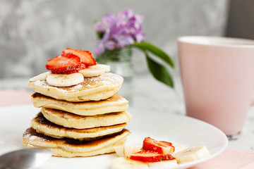 A stack of pancakes with a pink tea mug and  fresh strawberries on a white plate on a light background. A small bouquet of lilacs in a glass vase stands next to the Breakfast.