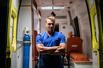 A handsome man in a medical uniform looking at the camera with a smile, arms folded, ambulance car in the background
