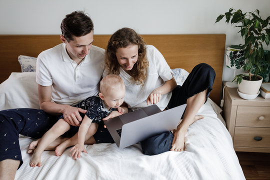 A Young Family Talk Via Video Call On A Computer In A Stylish Apartment On The Bed During Epidemic Covid. Mom, Dad And Son.