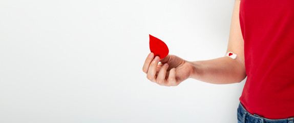 Blood donorship. Young girl in red T-shirt holds drop in her hand, the second hand taped with patch   with red heart after giving blood on gray background. Copy space