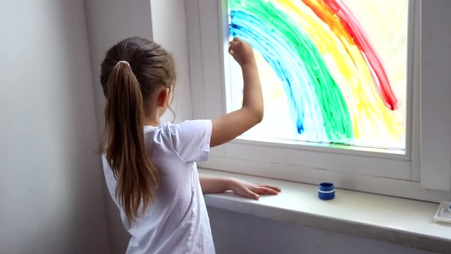 Little caucasian girl graws a rainbow on the window during coronavirus quarantine at home. Chase the rainbow flashmob