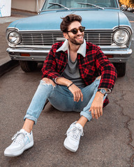 Handsome man wearing plaid red jacket and sunglasses, sitting on the street, in front of vintage car. - 349015602
