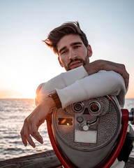 Portrait of a handsome man resting on a telescope. Fashion model standing by the ocean with his...