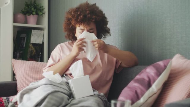 Sick african american girl with flu virus on sofa at home. She uses wipes to sneeze