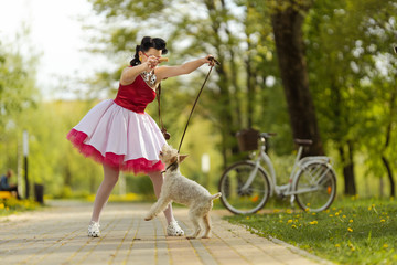 Obraz na płótnie Canvas A girl in a dress and hairstyle in the style of the 40-50s plays in the park with a dog breed Fox Terrier on a leash on a sunny day. Retro style photo. 
