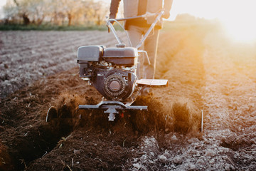 male farmer with a walk-behind tractor cultivates the land with a plow at sunset.