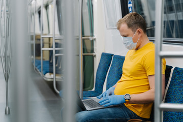 Fototapeta na wymiar Young man wears respiratory mask to prevent coronavirus, works freelance on laptop computer, poses in metro carriage, afraids of getting sick, travels in public transport. 2019-ncov. Keeping safe