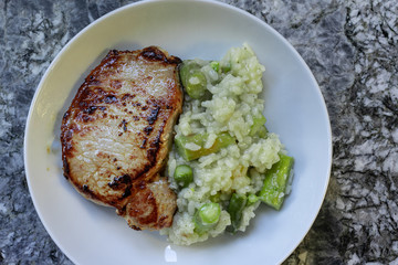 Roasted meat and risotto rice with asparagus in white round plate on granite background. High angle view.
