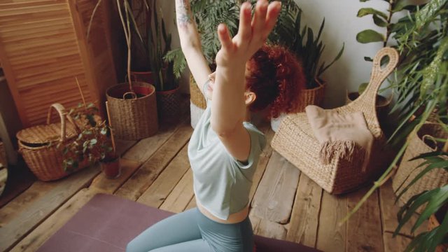 Arc shot of young beautiful woman sitting on the floor in room with green plants, raising arms, closing eyes and holding palms in prayer position in front of her chest while practicing yoga at home