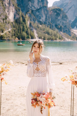 Fototapeta na wymiar Beautiful bride in a white dress with sleeves and lace, with a yellow autumn bouquet on background of the arch for ceremony, at Lago di Braies in Italy. Destination wedding in Europe, on Braies lake.