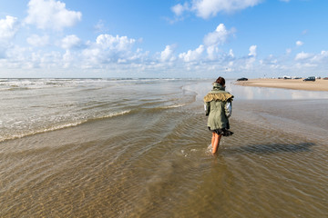 A woman walks along the clean, transparent waters of Romo Beach, Denmark,  wearing a green coat with some cars on the sand in the background. Beach concept