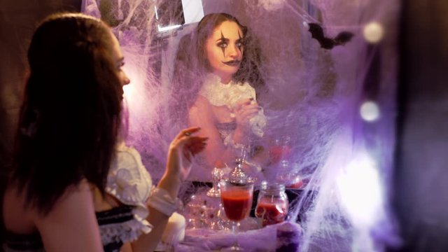 Portrait of a woman. Girl looks in the mirror. The mirror is decorated with cobwebs and bats. Model with makeup for Halloween. Woman flirting. The man raises a glass of red drink.