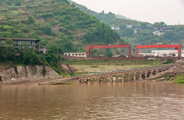 Naklejka premium Myaoyinan, Chongqing, China - May 8, 2010: Yangtze River. Slipway and large crane bridges behind brown water, at Chiandong Shipyard. Green Mountain slope with agriculture plots in back.