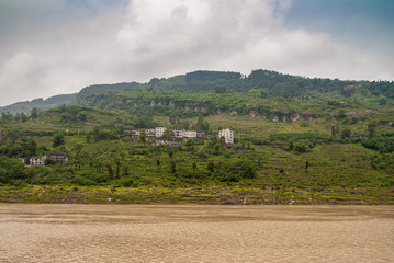Fototapeta na wymiar Fengdu, Chongqing, China - May 8, 2010: Yangtze River. Green hilly shoreline with agriculture plots in terraces and village with gray and whitish housing under blue cloudscape behind brown water.
