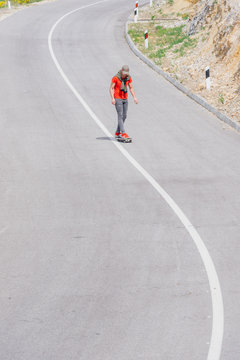 Male caucasian longboarder riding downhill on an empty road, preparing for a speed tuck on the next turn he has to make. Wearing a red t-shirt green hat and black jeans.