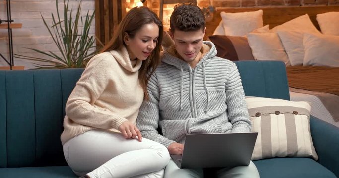 Young beautiful Couple having Fun while staring at Laptop, watching Comedy. Young Family is spending cozy, happy Evening at Home, wearing home Clothes. Having big Loft Flat-Studio. Lovers. Emotions.