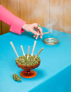 Hand in Pink Sleeve Holding Smoking Joint with Dish Full of Cannabis and Joints in Foreground on Blue Tablecloth