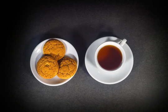 White cup with black tea and oatmeal cookies on a white saucer.