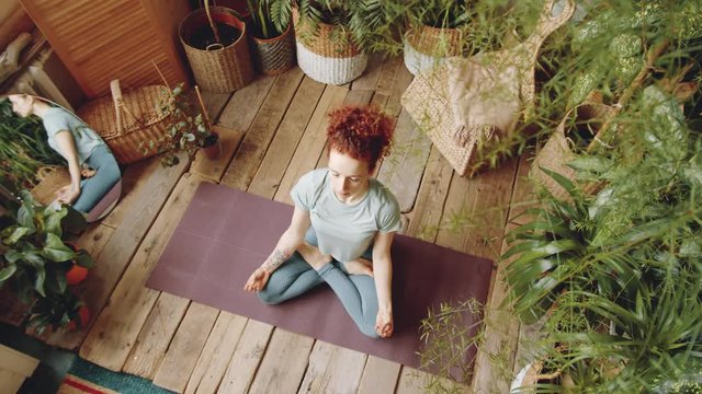 Top view of young redhead woman in sportswear sitting in lotus pose on yoga mat and holding hands in mudras while meditating at home in room filled with green plants