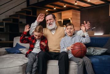 Obraz premium Concentrated senior bearded grandpa with his happy grandsons spending their leisure at revision the basketball championship on tv at home.