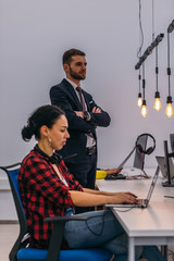 Picture of a handsome young manager standing next to his female coworker while she is working on the laptop