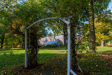 A wooden archway covered in vines sits in the foreground with a greenhouse attached to a large brick building in the background. The garden has a number of large mature trees with vibrant green grass.