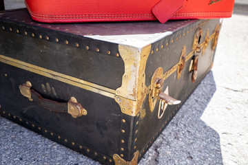 A vintage travel trunk with a blue exterior, brass edging and lock mechanisms. The suitcase is unlocked while sitting on a sidewalk. There's a bright red suitcase on top of the antique container. 