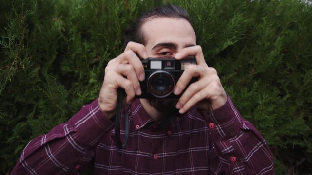 Hipster long-haired bearded young man takes pictures with old camera. Taking pictures towards the camera.