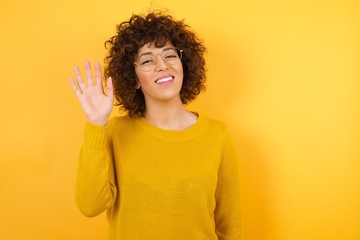Young beautiful brunette woman wearing casual clothes over yellow isolated background Waiving saying hello happy and smiling, friendly welcome gesture. © Roquillo