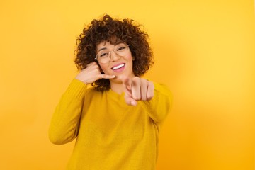 Fototapeta na wymiar young pretty brunette woman wearing yellow sweater smiling cheerfully and pointing to camera while making a call you later gesture, talking on phone against flat yellow color wall