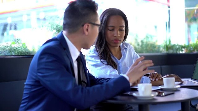 Two young business colleagues in formal wear discussing mobile application on smartphone during meeting in cafe.Asian entrepreneur together with African American secretary talking about web page
