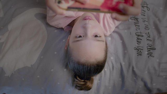 Little girl cute plays in smartphone lying on the bed
