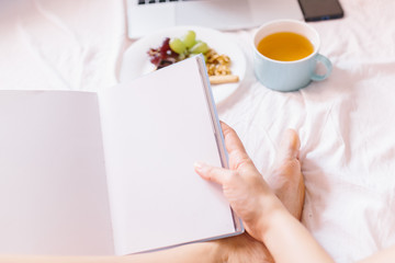 Young woman reading a book. Tea at home in her bed, checking her laptop, reading a book and having breakfast.