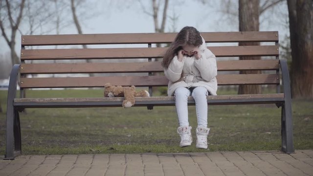 Crying lost little girl rubbing eyes as sitting on bench in spring or autumn park. Portrait of pretty lonely Caucasian kid with teddy bear. Childhood, sadness, social problem, anxiety.
