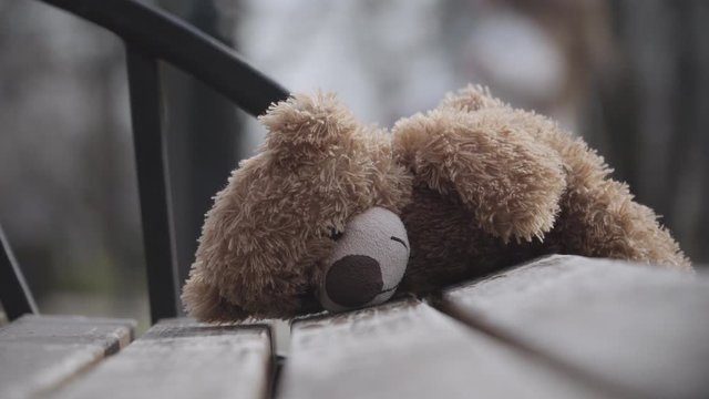 Brown teddy bear lying on bench with blurred curly-haired girl having fun at the background. Close-up of abandoned toy. Childhood, leisure, growth, lifestyle.