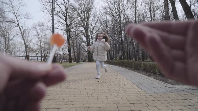 Kidnapper's point of view of little pretty Caucasian girl resting in city park alone. Blurred male hands holding lollypop and waving to cute brunette Caucasian child. Kid running to man and leaving.