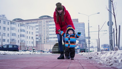 A young woman leads a infant by the hands down the street in winter. First steps baby