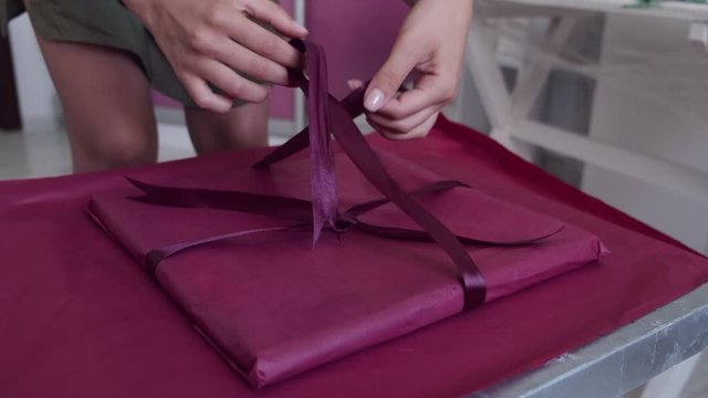 Person is wrapping gift with pink paper. Woman decoraes gift. Pink ribbon is used for making bow.