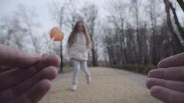 Little brunette Caucasian girl walking to kidnapper and taking lollypop. Criminal's point of view. Maniac taking hostage in spring or autumn park. Danger, social problem, kidnapping, crime.