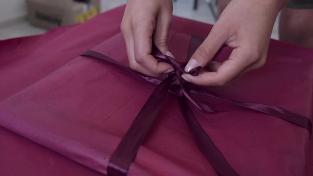 Person is wrapping gift with pink paper. Woman decoraes gift. Pink ribbon is used for making bow.