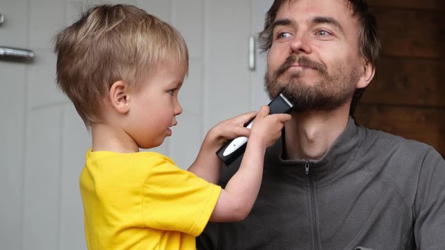 Little child boy cut father beard with clipper. Family haircut, hygiene, beauty and selfcare at home during quarantine lockdown, lifestyle concept.