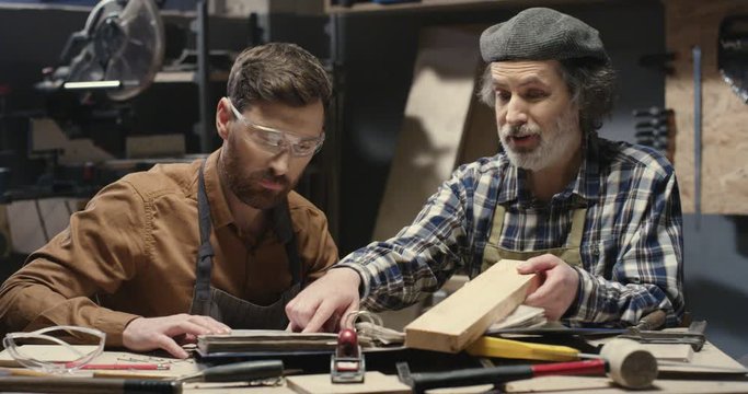 Older and younger Caucasian carpenters sittiing at table in workshop and talking. Male joiners, father and son, working with lumber in studio. Man teaching carpentry craft.