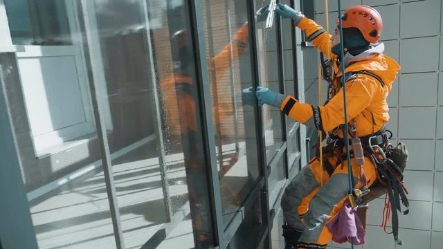 glazing, a young equipped industrial climber in an orange jumpsuit and helmet neatly washes the windows outside the building hanging on ropes, slow motion