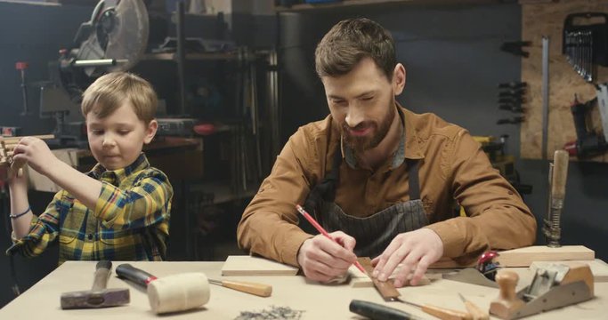 Cheerful handsome young Caucasian carpenter working at desk in workshop and small son playing next to him. Faher woodman drawing and measuring wood at table with little boy helping.