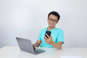 Asian young man happy and smile when he see on smart phone with laptop beside it. Indonesian man wearing blue shirt.