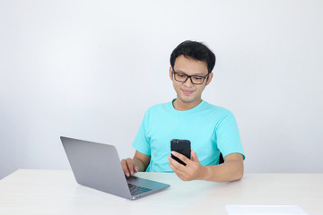 Asian young man happy and smile when he see on smart phone with laptop beside it. Indonesian man wearing blue shirt.