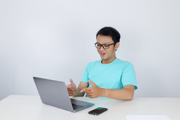 Young Asian man feeling happy and smile when work laptop on table. Indonesian man wearing blue shirt.