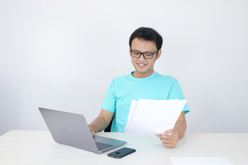 Fototapeta na wymiar Young Asian Man is smile and happy when working on a laptop and document on hand. Indonesian man wearing blue shirt.