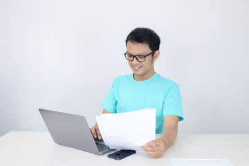 Young Asian Man is smile and happy when working on a laptop and document on hand. Indonesian man wearing blue shirt.